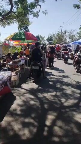 A busy outdoor market is depicted on a sunny day. Several vendors have set up stalls under colorful umbrellas, displaying goods. Pedestrians walk along a paved road, which appears to be a street serving as the market. Motorcyclists are also present, with some riding through the market and others parked. The ground is covered in dappled shadows from trees overhead. The overall atmosphere suggests a lively and active commercial gathering.
