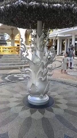 The video shows a view of a large, cone-shaped structure in an outdoor courtyard. The structure is silver and appears to be made of metal, with a flame-like design at the base and a canopy of numerous small, metallic leaf-shaped ornaments hanging from it. A circular, patterned pavement is visible beneath the structure, and a portion of a building with white pillars and a golden railing is in the background. Two adults and two children are present in the courtyard, standing a short distance from the structure. One child is wearing a striped dress, and the other is wearing shorts and a t-shirt. The adult with the children is holding a camera. The scene is well-lit, suggesting daytime.