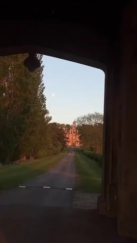 The video captures a serene view looking out from beneath a dark stone archway, possibly a gatehouse entrance to an estate. A long, paved driveway stretches directly ahead, flanked on both sides by meticulously maintained green lawns and tall, mature trees, some with prominent vertical forms, possibly poplars. In the far distance, a large, ornate building, appearing to be a historic stately home or castle, is visible, partially bathed in a soft, warm light, suggesting either dawn or dusk. Above the distant building, a bright, full moon is clearly visible against a pale blue, clear sky. The overall scene conveys a sense of grandeur and tranquility, characteristic of a large, well-preserved property in a rural setting within South Kesteven, United Kingdom. No people or specific activities are visible, and there is no discernible text.