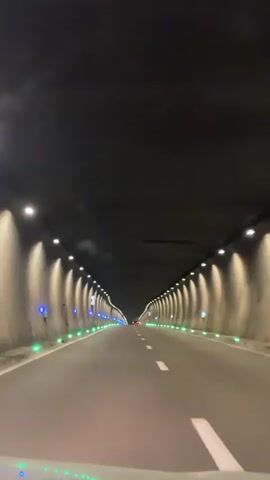 The video shows a first-person perspective of driving through a dimly lit tunnel. The tunnel has a dark ceiling and concrete walls illuminated by rows of upward-facing lights that cast a warm glow. Along the sides of the road, there are green and blue lights embedded in the pavement or walls. The road is paved and has white lane markings. The tunnel appears to be quite long, with the vanishing point of the road in the distance suggesting a significant length. There are no other vehicles clearly visible in the immediate vicinity, though faint red lights can be seen far ahead. The overall atmosphere is dark and enclosed, typical of a highway tunnel.