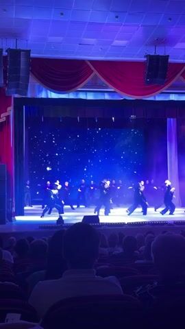 A wide shot shows a dance performance on a stage in a dimly lit auditorium. The stage features a large blue backdrop with numerous small white lights resembling stars, framed by red curtains on either side and a red valance at the top. Numerous performers, dressed predominantly in black attire, are dancing in synchronized movements across the stage, illuminated by bright blue and white spotlights. Some smoke effects are visible on the right side of the stage. Two large speaker arrays are suspended from the ceiling on both the left and right sides of the stage. The backs of several audience members seated in red chairs are visible in the foreground, watching the performance. Upbeat electronic music plays throughout the video.