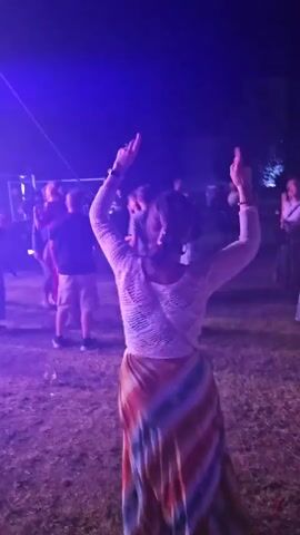 The video shows a group of people dancing at an outdoor event at night. Purple lighting illuminates the scene. In the foreground, a woman with her back to the camera is dancing, wearing a white crocheted top and a long, multi-colored skirt. She has her hands raised. Behind her, other people are dancing in front of a stage area with fencing. The ground appears to be covered in grass and dirt.