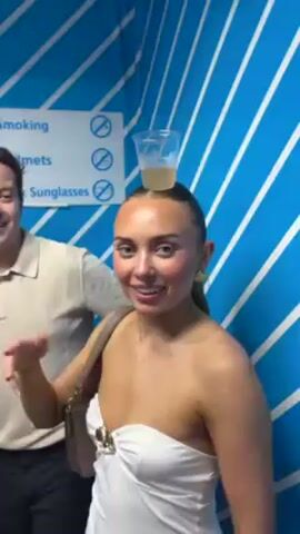 The video depicts a young woman with her hair pulled back and wearing a white strapless top, smiling directly at the camera. She has a small, clear plastic cup filled with a yellow liquid balanced on her head. She gestures with her left hand towards the cup while smiling. Behind her and slightly to the left is a man in a beige polo shirt and dark pants, partially visible. The background is a wall with a bright blue and white diagonal stripe pattern. There is also a white sign with black text and prohibition symbols indicating that smoking, helmets, and dark sunglasses are not allowed. The video appears to be taken in a confined space.