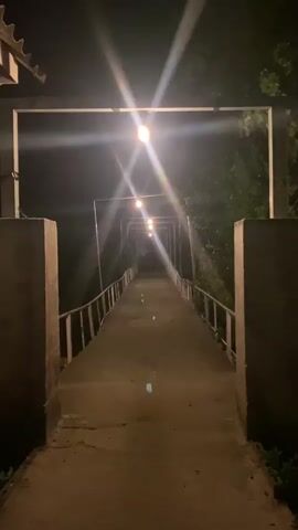 The video shows a concrete bridge at night, illuminated by several equidistant lights overhead. The bridge has white railings along each side. The lights have a bright, starburst effect on the camera. The environment is dark with trees visible on the right side of the frame. The camera angle is at the entry of the bridge, facing straight down the length of it.