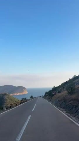 The video shows a view from a vehicle driving on a winding coastal road. The road has two lanes with a solid white line dividing them and a broken white line on the outer edge. The right side of the road is a steep, rocky hillside covered in dry grass and sparse vegetation. On the left side of the road, there is a stone retaining wall and the terrain slopes down towards the sea. In the distance, a body of water, likely the ocean, is visible, with a peninsula or large island extending into it. The sky is clear and blue with a few wispy clouds near the horizon. There are a few signs visible along the road, including a speed limit sign showing "40" and a warning sign indicating a sharp curve. The sun appears to be setting or rising, casting a soft light on the landscape. The vehicle appears to be the only one visible on the road in the immediate vicinity.