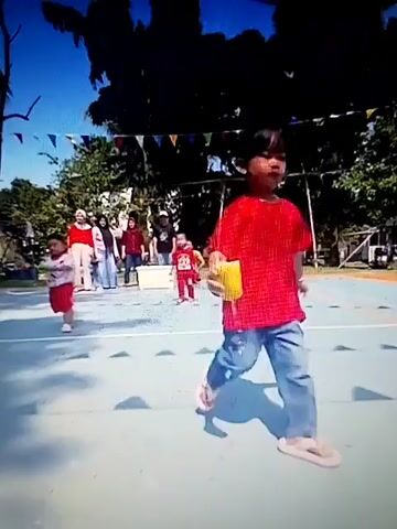 A low-angle shot shows a young child in a red shirt and pants running towards the camera on an outdoor court with a blue and green surface. The child is smiling and holding a yellow object. In the background, several people, including adults and children, stand watching. A string of triangular flags is strung across the top of the frame. A hand enters the frame from the right, reaching out towards the child.
