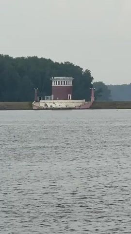 A video shows a body of water with a structure resembling a boat with a tower on it in the distance. The water is choppy, suggesting a slight breeze. Trees line the horizon behind the structure. The sky is overcast and a dull gray. The structure has a long, white hull and a red and white tower on top. The video is very shaky and the image quality is poor.