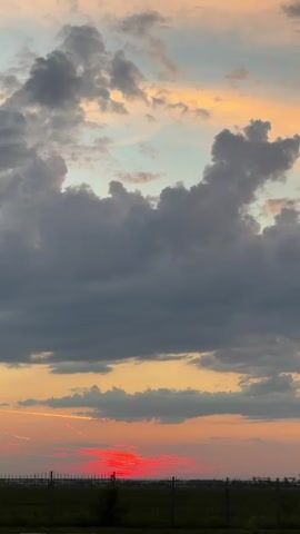 The video captures a sunset with vibrant colors painting the sky. The lower portion of the frame shows a fence and some vegetation, suggesting an outdoor setting like a field or park. Above, dramatic clouds are illuminated by the setting sun, creating a beautiful contrast between the dark gray clouds and the warm hues of orange and red. The overall scene is serene and picturesque, showcasing the natural beauty of the sky at dusk.