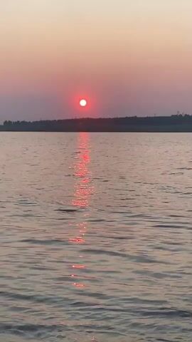 The video shows a body of water with gentle waves reflecting the setting sun. The sun is a bright red orb low in the sky, casting a shimmering path of light across the water. The horizon is lined with a dark silhouette of land, possibly trees or hills. The sky is a soft gradient of pink and orange, indicating the transition from day to night. The overall scene is serene and picturesque, typical of a sunset over a lake or river.