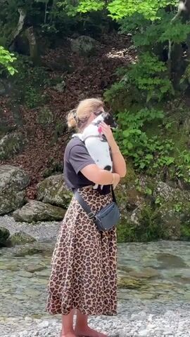 A woman with blonde hair tied back in a bun stands barefoot by a shallow stream. She is wearing a black t-shirt and a long, leopard-print skirt. She holds a small black and white dog, possibly a Jack Russell Terrier, cradled in her arms. The dog is wearing a red collar. A small black crossbody bag is slung across the woman's chest. The background features lush green trees, rocks, and a dirt path. The woman and dog are facing away from the camera, looking towards the trees and rocks. The water is clear and shallow, with visible rocks and pebbles on the stream bed. Sunlight filters through the trees, creating dappled light on the scene. The overall atmosphere is tranquil and natural.
