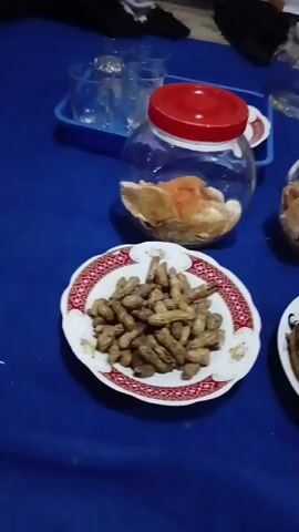 A close-up, slightly shaky video shows a table covered with a blue cloth. On the table, there are two clear glass jars with red lids filled with snacks. In front of the jars are two plates. The plate on the left holds a pile of peanuts in their shells. The plate on the right holds several cooked bananas, some of which are dark and appear overripe. To the right of the plate of bananas is a silver teapot. In the background, there are a few clear glasses, one on a blue tray, and a person's legs and feet are visible, indicating a seated individual. The lighting is dim, suggesting an indoor setting.