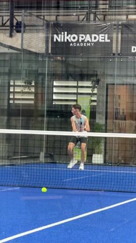 A young man is playing padel. He is wearing a grey tank top and black shorts. He is holding a blue and black padel racket. The court is blue with white lines. There is a net across the middle of the court. There is a ball in the air that he is about to hit. There are glass walls around the court. There is a banner in the background that says "Niko Padel Academy".