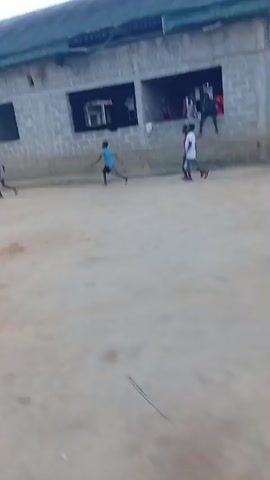The video shows a group of people gathered on a dirt field. They appear to be involved in some sort of game or activity, as they are moving around and interacting with each other. Some individuals are wearing sports clothing. In the background, there's a building with windows and a roof, and trees are visible in the distance. The sky is overcast, suggesting it may be early morning or late afternoon.