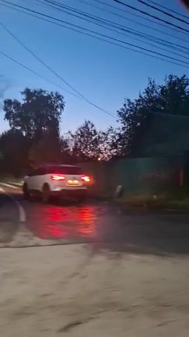 The video shows a dimly lit outdoor scene at dusk or dawn. A white car with illuminated red taillights is visible on a wet road, suggesting recent rain. Two figures, one in a white outfit and the other in a yellow and white outfit, are walking on the side of the road. Behind them, houses and trees line the street, with power lines visible overhead against the twilight sky. The overall atmosphere is calm and quiet.