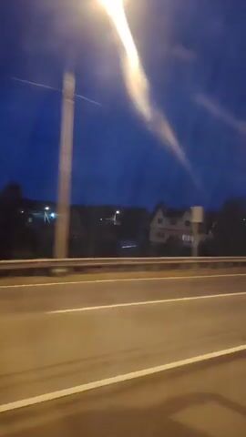A short video clip shows a view from a moving vehicle at dusk or night. The sky is a deep blue, and there are some faint streaks of light, possibly from passing cars or clouds. In the middle ground, a two-story house with illuminated windows is visible, nestled among trees. A highway with white lane markings stretches across the foreground, appearing wet and reflecting light. A metal guardrail separates the highway from the landscape beyond. A lamppost with a bright light is also visible on the right side of the frame. The overall impression is one of a serene, yet somewhat isolated, rural or suburban setting experienced from the perspective of someone traveling on a road.