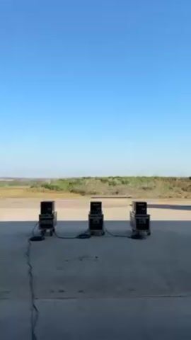 The video shows three black rectangular objects, each stacked on two smaller black boxes on wheels. These are arranged in a horizontal line on a concrete surface, connected by black cords. Behind them is a field of green and brown vegetation under a clear blue sky. Suddenly, flames erupt from the top of each stack, shooting upwards and outwards in fiery orange bursts. The flames quickly dissipate, leaving trails of smoke.