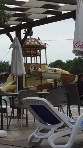 The video shows a water slide structure with slides. In the foreground, there are empty lounge chairs and umbrellas by a pool. A few people are visible in the background, some sitting and some near the pool. The weather appears overcast.