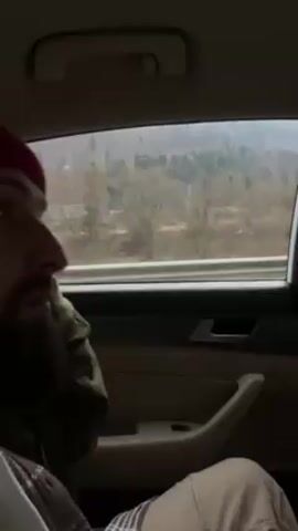 The video depicts a man with a beard and a red beanie hat, sitting in the back seat of a car. He looks out the window at a landscape featuring mountains, trees, and some snow. The camera is positioned inside the car, focusing on the man and the view outside the window. The car's interior is visible, including the door panel and a handle. The overall atmosphere suggests a journey or travel through a mountainous region.
