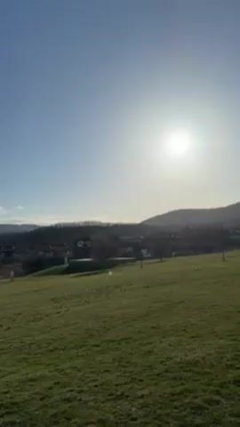 The video shows a sunny, daytime landscape with a green grassy field in the foreground. In the background, there's a small village or group of buildings, and behind that, rolling hills and mountains are visible under a bright blue sky. The sun is shining brightly, creating a slight haze in the distance.