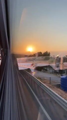 The video captures a sunset viewed from inside a vehicle, likely a bus, given the prominent reflection of the vehicle's interior on the left side of the frame. The sun, a bright orb, is low on the horizon, casting a warm orange glow across the sky and landscape. The scene appears to be a roadside environment, possibly a highway or a rural road, with a paved road stretching into the distance. Guardrails line the side of the road, and there are signs, including a stop sign. Vegetation, including trees and bushes, dots the landscape, and there are some buildings or structures visible in the background. The light suggests either sunrise or sunset, with the sun’s rays illuminating the clouds and hills. The camera's perspective indicates movement, suggesting the vehicle is in motion.