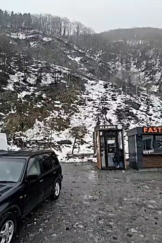 The video shows a snowy mountain landscape with a black SUV parked in front of two food stands. The camera then moves to reveal a crowded scene of cars at the Upper Lars border crossing. The road is wet, and snow is falling. There are many cars of various colors and sizes, including a blue van. Concrete barriers line the side of the road. A multi-story building can be seen in the background.
