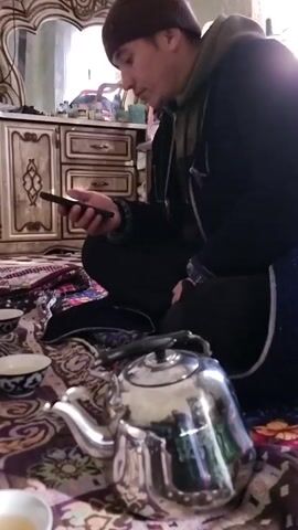 Here is a concise description of the video:

The video shows two men sitting on a rug in a room. One man is looking at his phone and the other man is talking, gesturing with his hands. There is a teapot and small cups on the rug, along with some round flatbread. The room appears to be a bedroom with a dresser and a doorway visible. The lighting in the room is somewhat dim.