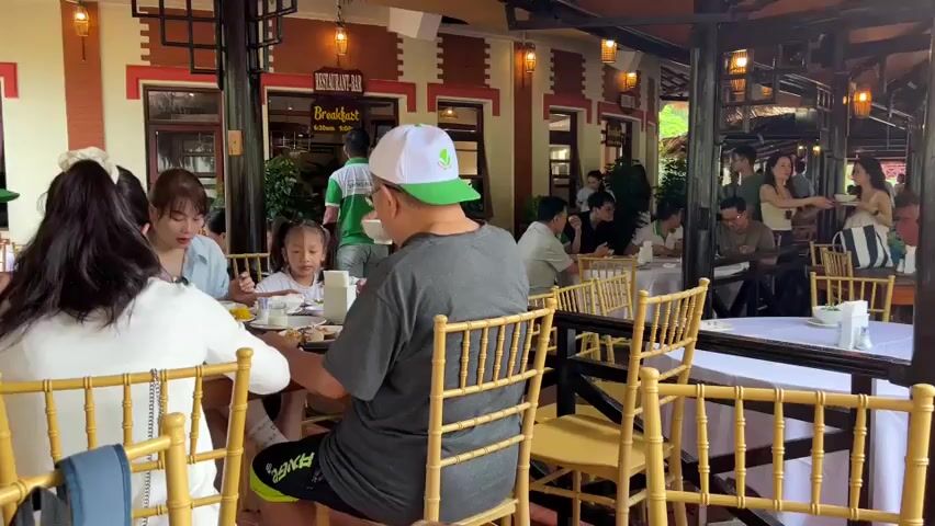 The video shows a busy restaurant interior with many patrons seated at tables. In the foreground, a man in a gray t-shirt and a white and green baseball cap is seated at a table facing away from the camera. To his left, a woman with dark hair and a young girl are also seated at the same table. The restaurant appears to have a traditional architectural style with wooden beams and decorative lighting. Several other tables are occupied by groups of people eating and conversing. In the background, more tables and patrons are visible, as well as a doorway labeled "BREAKFAST." The overall atmosphere suggests a lively dining establishment.