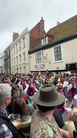 The video shows a crowd of people watching a traditional dance performance in a street lined with buildings.  The dancers are wearing colorful vests and flower crowns. Some people in the crowd are recording the performance with their phones, while others are holding drinks. There is a shop called "Through the Looking Glass" in the background. The buildings are mostly white with dark windows and some have red brick detailing. The sky is overcast.