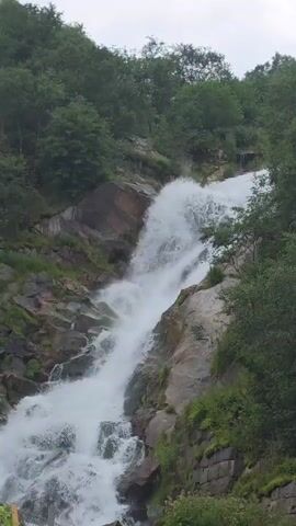 A powerful waterfall cascades down a rocky mountainside covered in lush green trees. The water is a foamy white as it plunges over the rocks, creating a mist that rises into the air. The sky above is overcast, lending a soft, diffused light to the scene. The overall impression is one of raw natural beauty and untamed power.