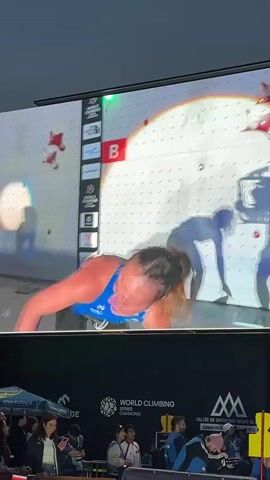 A woman in a blue tank top and black pants is projected onto a large screen. She is on a climbing wall with red holds. She is looking to her left and smiling. In the background, another climber is projected onto the screen. Below the screen, there is a crowd of people watching. There are tents and banners that say "WORLD CLIMBING" and "CHAMONIX." There are also stage lights.