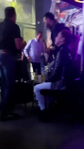 The video shows a group of people in what appears to be a bar or nightclub. There are two men in black clothing, possibly security or staff, flanking a man in a light blue shirt who is dancing or moving energetically. A woman with a ponytail is seated nearby, drinking from a glass. There are drinks on the table in front of her. The lighting is dim and the background features a wall with signage. The general atmosphere seems lively and social.