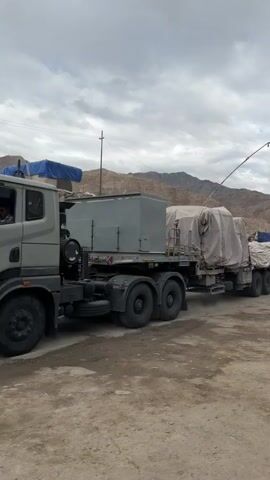 The video shows a convoy of trucks on a dusty road with mountains in the background. The trucks are large and appear to be transporting heavy equipment, as evidenced by the tarps covering the cargo. There is a man walking in the foreground, looking towards the trucks. In the background, another truck is visible, as well as a sign that reads "LADAKH POLICE DISTRICT POLICE LEH". The sky is overcast with clouds. The overall impression is of a logistical operation in a remote or mountainous region.