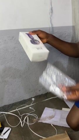 A person with dark skin is shown from the knees up. They are wearing blue shorts. They are kneeling on a floor with a gray concrete wall behind them. They open a white box labeled "F015." The floor has various items, including white wires, a white phone, a yellow adapter, some paper, and a cardboard box. The person pulls a small, white, handheld fan out of the box. The fan has a copper-colored band at the top and bottom and a white base.