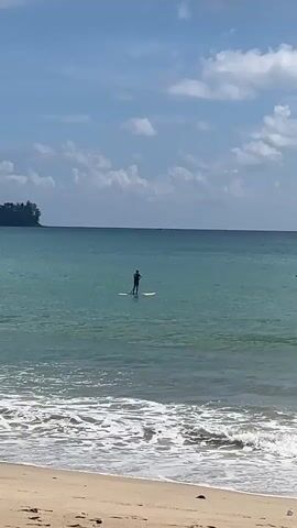 A person is paddleboarding in the ocean. The water is turquoise and clear, with small waves lapping at the sandy shore. In the distance, a lush green hillside rises from the sea, dotted with buildings. The sky is a bright blue, with fluffy white clouds scattered across it. Another person is walking along the beach, facing away from the camera. The camera pans to the left, revealing more of the beach and the treeline.
