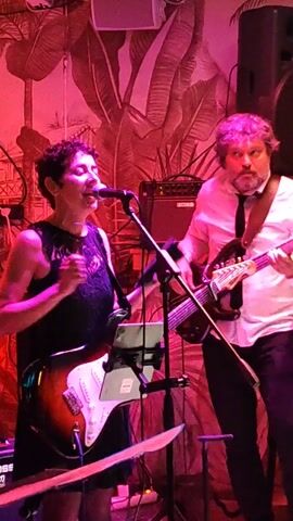 A woman with short dark hair wearing a black lace dress stands in front of a microphone and a guitar stand, singing and playing a sunburst electric guitar. To her right, a man in a white shirt and black tie plays a four-string bass. The background is a wall decorated with a faded print of palm trees and a vaguely visible cityscape, illuminated by red and purple stage lights. A black Boss amplifier is visible behind the woman.