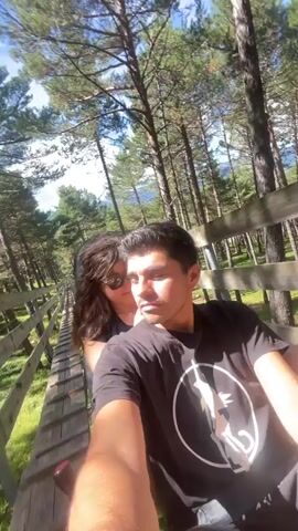 This is a POV video taken by a person with dark hair, wearing a black t-shirt with a white circular graphic and black shorts, seated in a green cart. Behind them, a person with long, wavy brown hair and sunglasses, wearing a black top, is also in the cart. They are on a luge track that winds through a forest with tall pine trees and a wooden railing. The track appears to be made of wood or a similar material with metal rails. The cart is in motion, moving down the track. The person in front is looking at the camera and appears to be speaking or reacting to the ride, while the person behind them is touching their hair and looking to the side. The lighting suggests it is daytime with bright sunlight filtering through the trees.