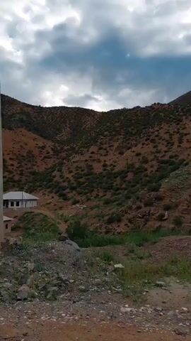 The video captures a scenic drive through a mountainous, rural landscape. The terrain is characterized by dry, rocky hillsides sparsely covered with low-lying green vegetation. Several simple, modern houses with white roofs and light-colored walls are nestled into the slopes and in a small valley. A dirt road, wet in places with visible puddles, winds through the scene. Tall, slender trees, possibly poplars, line portions of the road and fields.

In the foreground, a person wearing a dark jacket and pants stands near a small, fenced-in area with a yellow structure, possibly a greenhouse or storage unit. A car's side mirror is visible on the right side of the frame, indicating the perspective is from inside a vehicle. A tall, grey utility pole stands prominently near the center of the frame.

The sky is overcast with dramatic, heavy clouds, suggesting either early morning or late afternoon, and the possibility of recent or impending rain. The overall impression is one of a remote, agricultural community situated in a rugged natural environment. The activity appears to be a slow drive or passage through this area, allowing for observation of the landscape and its few structures. There is no visible text in the media file.