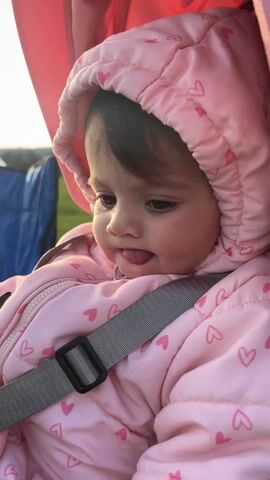 The video opens with a close-up of a baby in a pink hooded jacket with a heart print, sitting in a stroller and sticking their tongue out. The camera then pans to the right, revealing a scenic view of a lake at sunset. The sun is low in the sky, casting a warm glow on the water. There is a blue camping chair on the grassy bank. In the distance, a person is standing by the water's edge, casting a fishing line. The person is wearing a dark jacket and teal pants. The video appears to be taken at dusk.