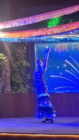 A woman in a patterned dress is performing a flamenco dance on a stage. The stage is lit with blue and purple lights. In the background, there is a banner that reads "4R Playa Paraiso" and is decorated with fireworks and confetti. Colorful streamers are strung across the top of the stage. The woman is in motion, with her arms raised and her skirt fanned out.