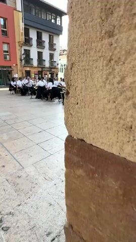 The video opens with a close-up of a young woman playing a saxophone. The camera then pans to the right, revealing a plaza with a band playing in the background. The band members are dressed in white shirts and black pants. Several people are seated in chairs in front of the band, listening to the music. The plaza is paved with stone tiles and surrounded by buildings of various colors. The sky is clear and blue. In the foreground, the woman continues to play her saxophone. The camera then moves to a wider shot of the plaza, showing more of the band and the audience. Some of the audience members are in wheelchairs. The video ends with a shot of the buildings surrounding the plaza.