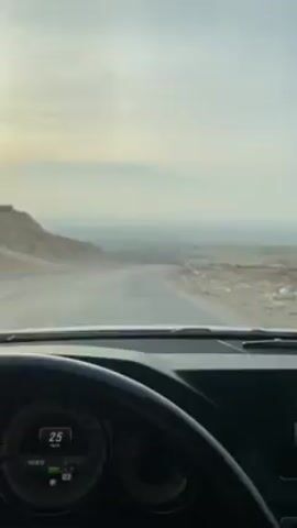 The video shows a first-person view from inside a car driving on a winding road. The landscape is arid and mountainous with low-lying vegetation. The sky is overcast and hazy. Another car is visible parked on the side of the road. The sun is visible on the horizon. The dashboard and steering wheel of the car are visible in the foreground. Loud music is playing. The car’s speedometer reads 22.