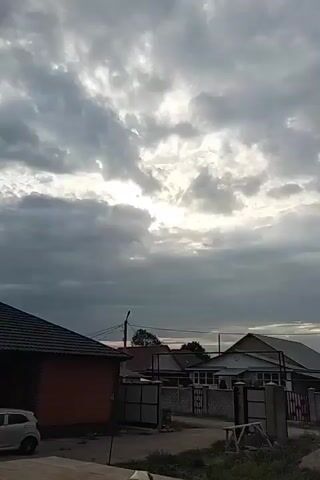 The video is a static shot of a cloudy sky above a few houses with fenced yards. The sun is visible behind the clouds, creating a bright area in the sky. The houses appear to be of a similar style and are in various states of construction or maintenance. There is a street or dirt path in front of the houses, and some construction materials are visible in the foreground. The overall impression is a rural or suburban setting under overcast skies.
