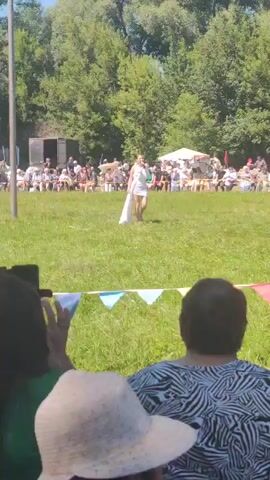 A female performer in a short white dress and a long white sash walks across a grassy field towards a seated audience. She holds a microphone in her right hand and appears to be singing or speaking. The audience is seated in rows of chairs on the grass, facing the performer. Behind the audience are trees and a blue tent. In the foreground, two spectators are visible from behind. One is wearing a green shirt and the other a black and white striped shirt. A third spectator in a straw hat is also visible. Colorful triangular flags are strung across the field in front of the audience. A tall pole stands to the left of the frame. The sky is blue and the sun is shining, indicating it is daytime.