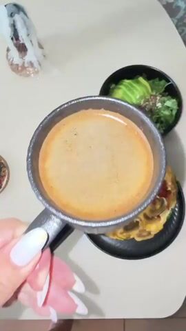 The media file shows a top-down view of a breakfast table setting. The main subjects are a black mug filled with dark coffee, a black plate with a slice of quiche or frittata topped with mushrooms and tomatoes, and a black bowl containing a salad with avocado, tomatoes, and greens. A gold fork rests on the plate. A hand with white-painted fingernails is holding the coffee mug. In the background, a decorative candle or sculptural object with white, wispy elements is visible, along with a small, round coaster with decorative text. The scene appears to be indoors, likely in a home, given the casual setting. The lighting suggests it could be morning or a well-lit indoor space. No specific weather or time of day is explicitly indicated beyond the breakfast context. The overall mood seems relaxed and leisurely, focused on enjoying a meal. The visible text on the coaster is difficult to read clearly but appears to be part of a decorative design. The location context provided is Oryol-Izumrud, Russia.