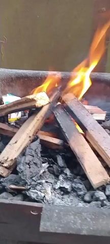 This is a close-up video of a small campfire being built and lit. The scene appears to be outdoors, possibly in a backyard or campground, with a neutral-colored wall in the background. The campfire is contained within a metal fire pit or grill.

The primary subjects are pieces of wood, some of which are already burning, and embers and ash at the base of the fire. In several frames, a person's hand is visible, interacting with the fire. This interaction involves adding more wood to the flames. The activity is the process of starting and tending to a fire.

Notable details include the bright orange and yellow flames, white smoke rising, and the rough texture of the burnt and unburnt wood. The time of day is not explicitly indicated, but the lighting suggests it could be daytime or early evening. There are no clear weather cues. The location is identified as Iskrets, Bulgaria. No text is visible in the video. The overall impression is one of a practical or recreational activity of fire-building.