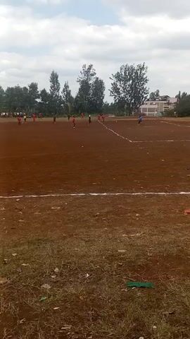 A distant view of a soccer game in progress on a dirt field, with a white line marking the near edge of the field. Multiple players in red uniforms are visible on the field, along with several players wearing dark colors, and a goalie in blue near the far goal line. Spectators stand along the sideline of the field, obscured by trees in the background. A building is also visible in the background. The sky is partially cloudy.