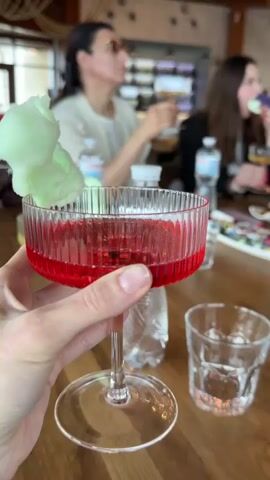 The video shows a group of women seated around a wooden table in a restaurant. The table is adorned with glasses filled with red and yellow liquids, water bottles, vases of flowers, and trays of small sweets. The camera focuses on a hand holding a glass of red liquid topped with green cotton candy. In the background, the other women are holding similar glasses and appear to be conversing. The restaurant has a rustic, yet elegant decor with hanging light fixtures and neutral-toned walls. A waiter can be seen in the background adjusting a chair.