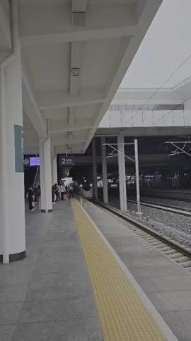 A high-speed train approaches a station platform. The platform has a gray tiled floor with a yellow tactile warning strip running along the edge. A white covered overhang supported by columns extends over the platform. On the left side of the platform, a white column stands prominently. Several people are waiting on the platform, some with luggage. A digital sign displaying "2" is visible overhead. The train, with a white and silver color scheme, is illuminated by its headlights as it moves along the railway tracks. The background shows more tracks and some elevated structures, suggesting an urban railway environment. The lighting indicates it is either dusk or a dimly lit indoor station.