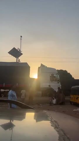 A busy street scene under a sky brightened by a setting sun. In the foreground, the distorted reflection of the setting sun appears on a car's hood. Several people mill about, including a man in a blue shirt and a group of women in brown robes. Behind them, a large truck covered with a dark tarp and a white semi-truck are parked.  A yellow tricycle, also known as a Keke Napep, pulls up next to the truck. A structure with solar panels is mounted on a pole on the side of the road. The scene looks like it is taking place in the late afternoon or early evening.