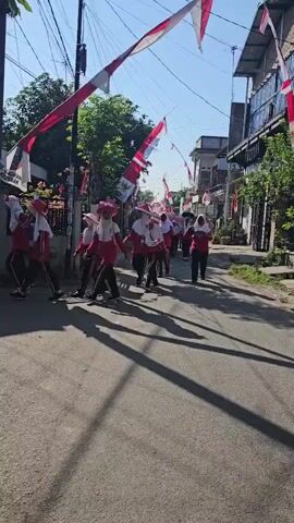 A group of children in red and white uniforms walk down a paved street lined with houses and trees. Indonesian flags are strung across the street, some flying from poles and others tied to buildings. The children appear to be participating in a parade or procession. The sun is shining, casting long shadows on the street. The children are walking in the same direction, towards the background of the image. The overall atmosphere appears festive and patriotic.